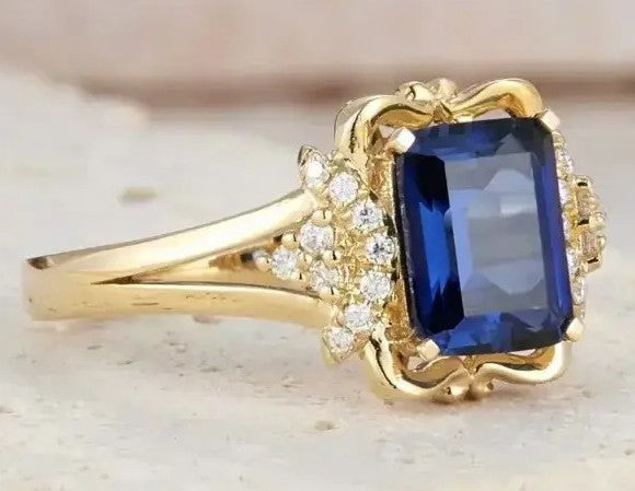 A yellow gold ring featuring a large rectangular blue sapphire gemstone, surrounded by small clear accent stones and an ornate decorative setting, shown on a light textured surface.