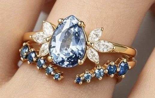 A gold ring with a large pear-shaped blue gemstone and smaller blue and clear accent stones, shown worn on a hand.