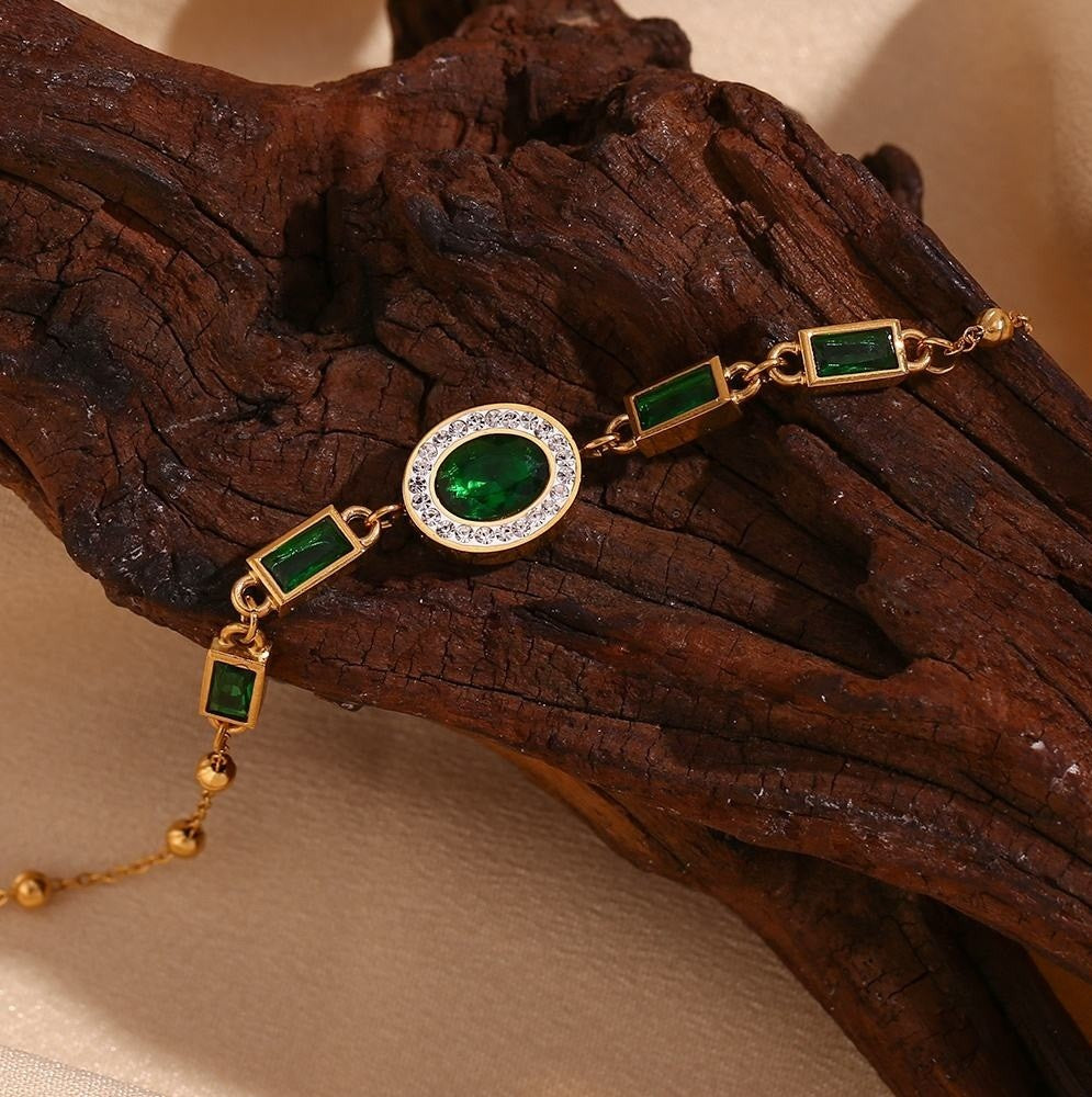 Gold-plated bracelet with emerald-cut green gemstones and a central oval green stone surrounded by clear crystals, displayed on a piece of dark wood.