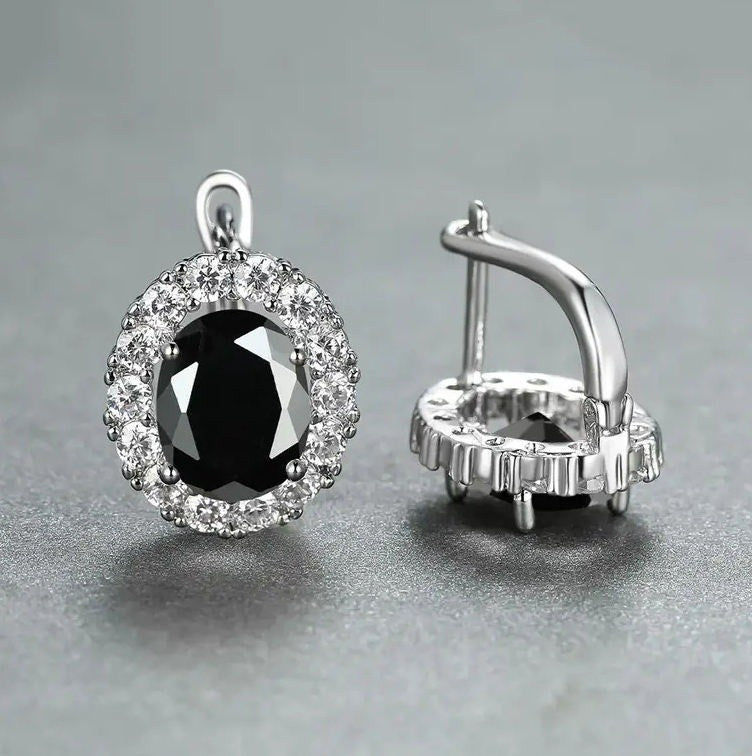 A pair of silver hoop earrings with a large oval black gemstone surrounded by a halo of small clear stones, shown on a neutral gray background.