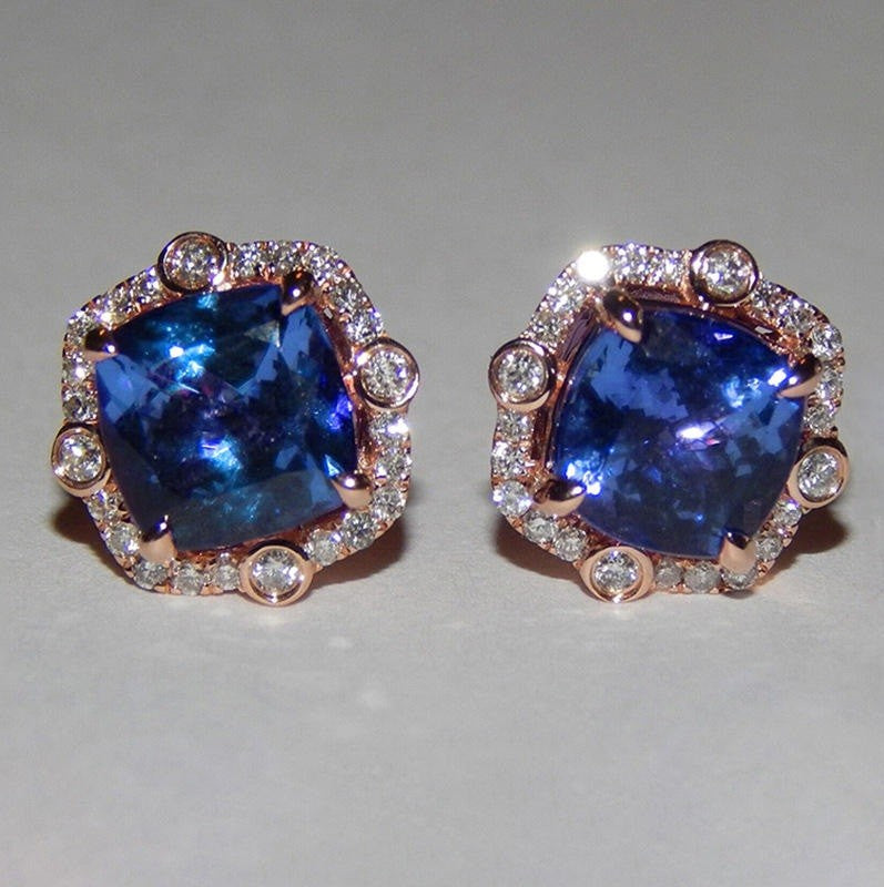 A pair of cushion-cut blue sapphire stud earrings set in rose gold with a halo of small clear stones, displayed on a neutral light gray background.