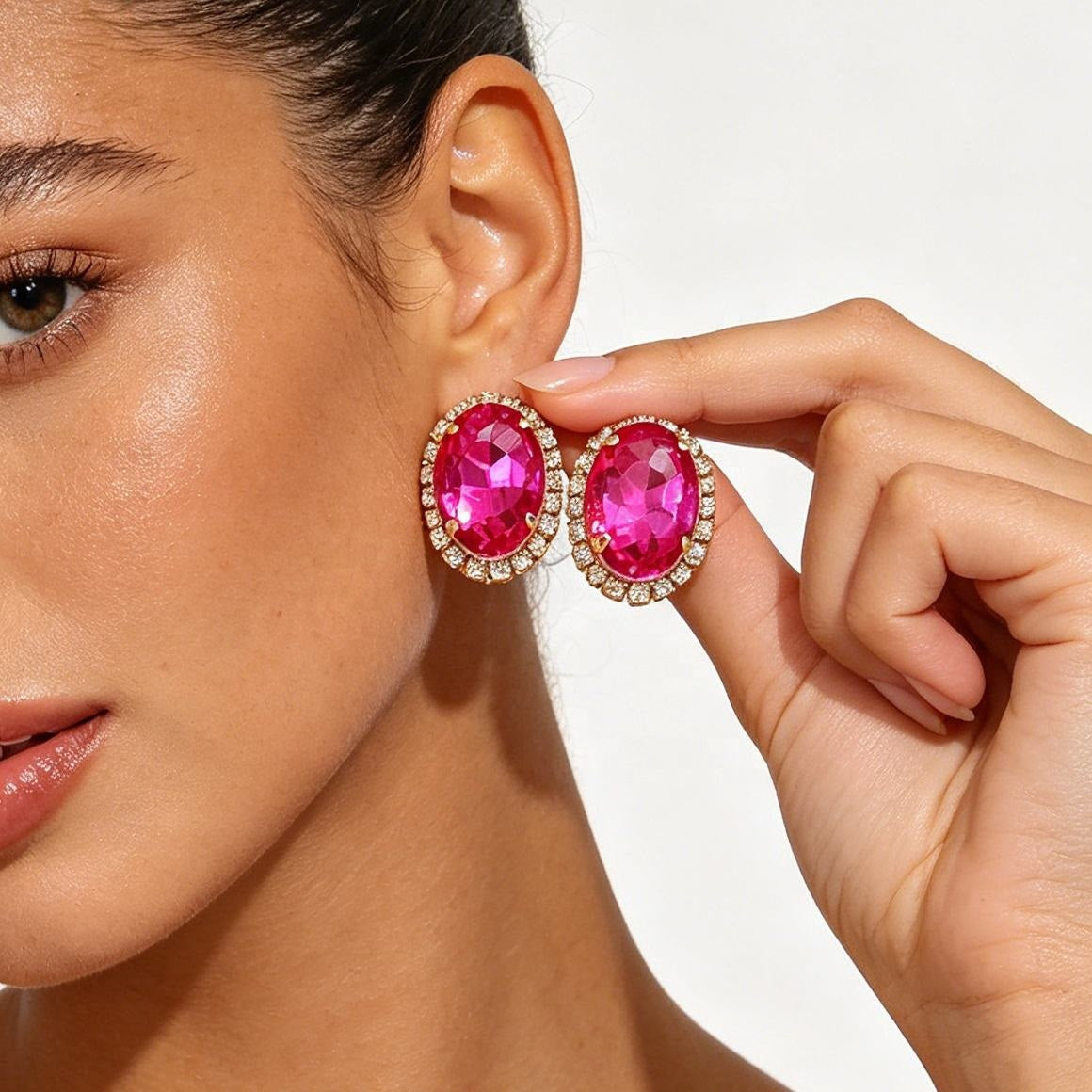 A close-up lifestyle shot of a model wearing oval pink gemstone stud earrings with gold halos and clear accent stones, while her hand holds the matching earring against a clean white background.