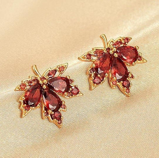 A pair of gold-toned leaf-shaped earrings with faceted dark red gemstones, displayed on a beige satin fabric background.
