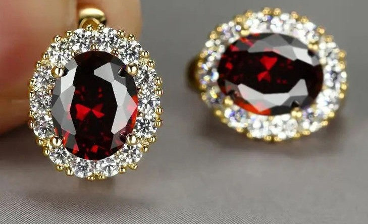 A pair of oval halo earrings featuring deep red faceted center stones surrounded by clear pavé-set stones in yellow gold settings, with one earring held against a finger and the other resting on a neutral gray surface.
