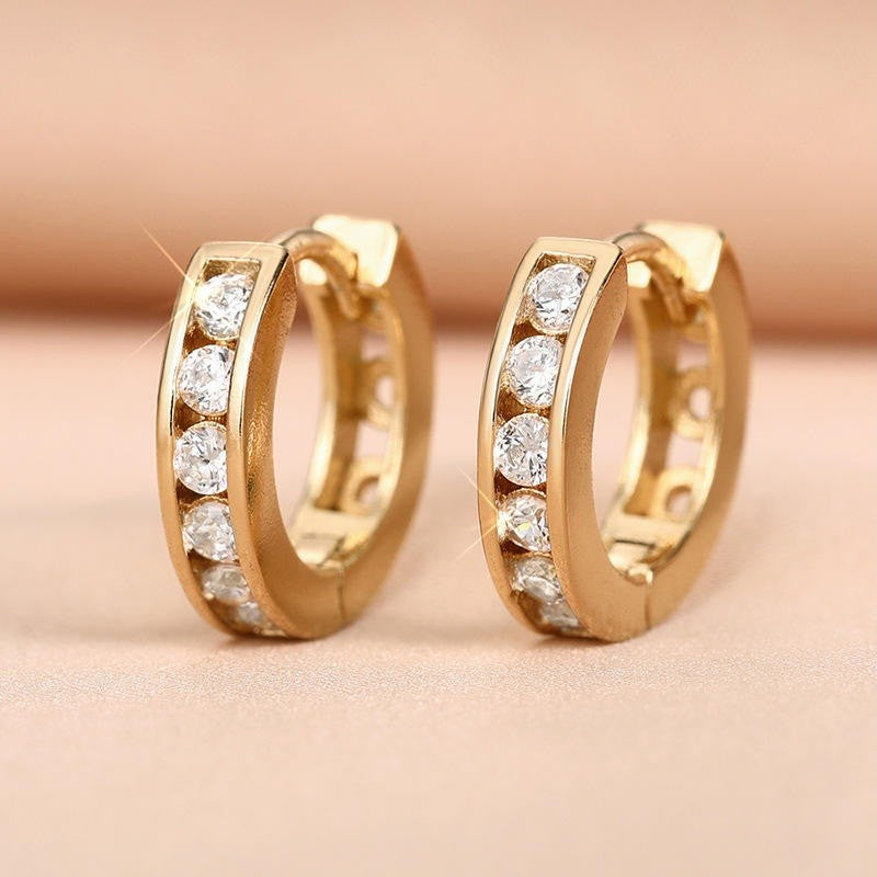 A pair of gold huggie hoop earrings with channel-set clear cubic zirconia stones, displayed on a soft beige surface.