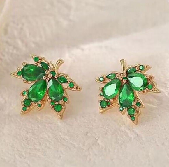 A pair of gold leaf-shaped stud earrings with green gemstones, displayed on a textured white surface.