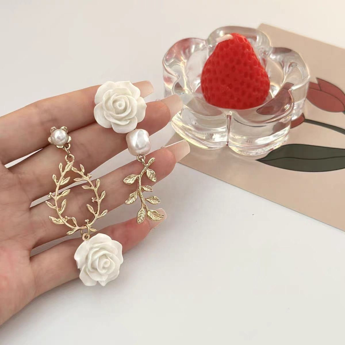 A hand holds a pair of dangling earrings with white rose charms, white pearl beads, and gold-toned leafy branches. A clear flower-shaped dish with a red strawberry sits on a beige card in the background.