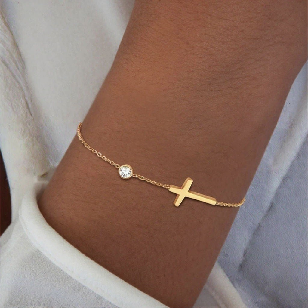 Gold bracelet with a cross and small round charm on a wrist.