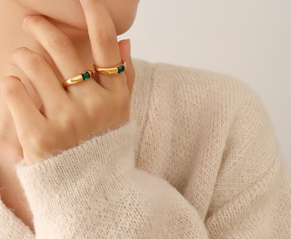 Close-up of a hand wearing two gold rings with green gemstones on a neutral background.
