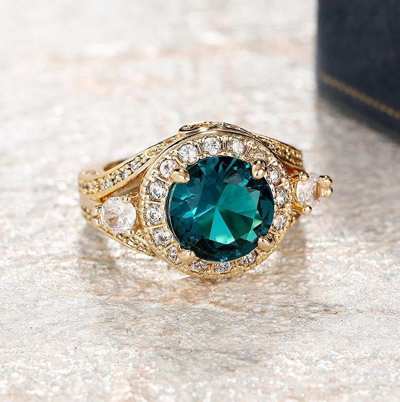 A gold ring featuring a large round faceted teal green center stone surrounded by a halo of small clear stones, with additional clear stones along the band, shown on a textured light surface.