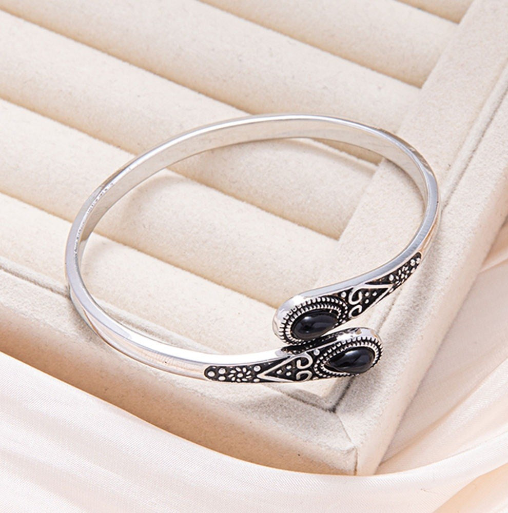 A silver-toned open cuff bangle bracelet with two black oval stones and ornate filigree detailing, displayed on a cream-colored jewelry box.