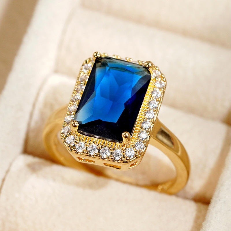 A gold ring featuring a rectangular blue gemstone surrounded by a halo of small clear stones, displayed on a cream-colored ring box.