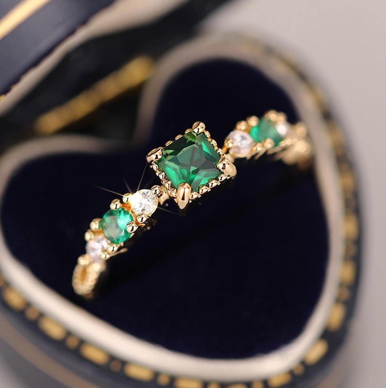 A gold ring with three emerald-cut green gemstones and small clear accent stones, displayed inside a heart-shaped dark blue ring box.