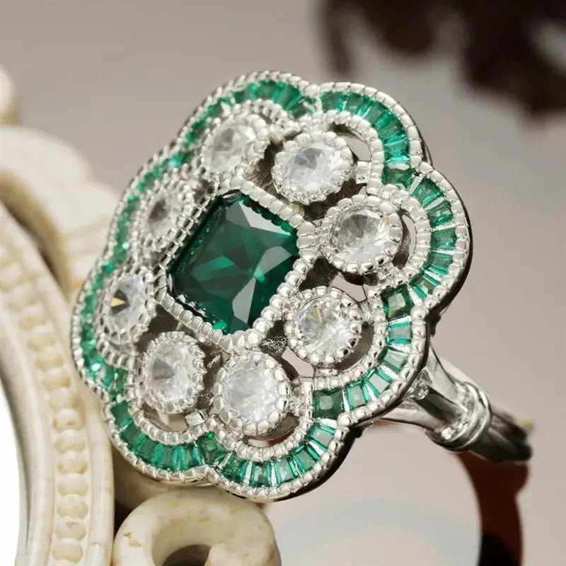 An ornate Art Deco style ring featuring a central emerald-cut green gemstone surrounded by a floral halo of round clear stones and baguette-cut green stones in a silver-toned setting.