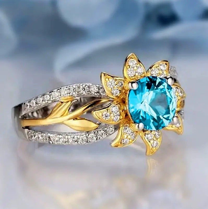 A two-tone ring featuring a large round-cut light blue gemstone in a gold flower-shaped setting, surrounded by small clear stones, with a split band combining yellow gold and silver-toned metal also paved with clear stones.