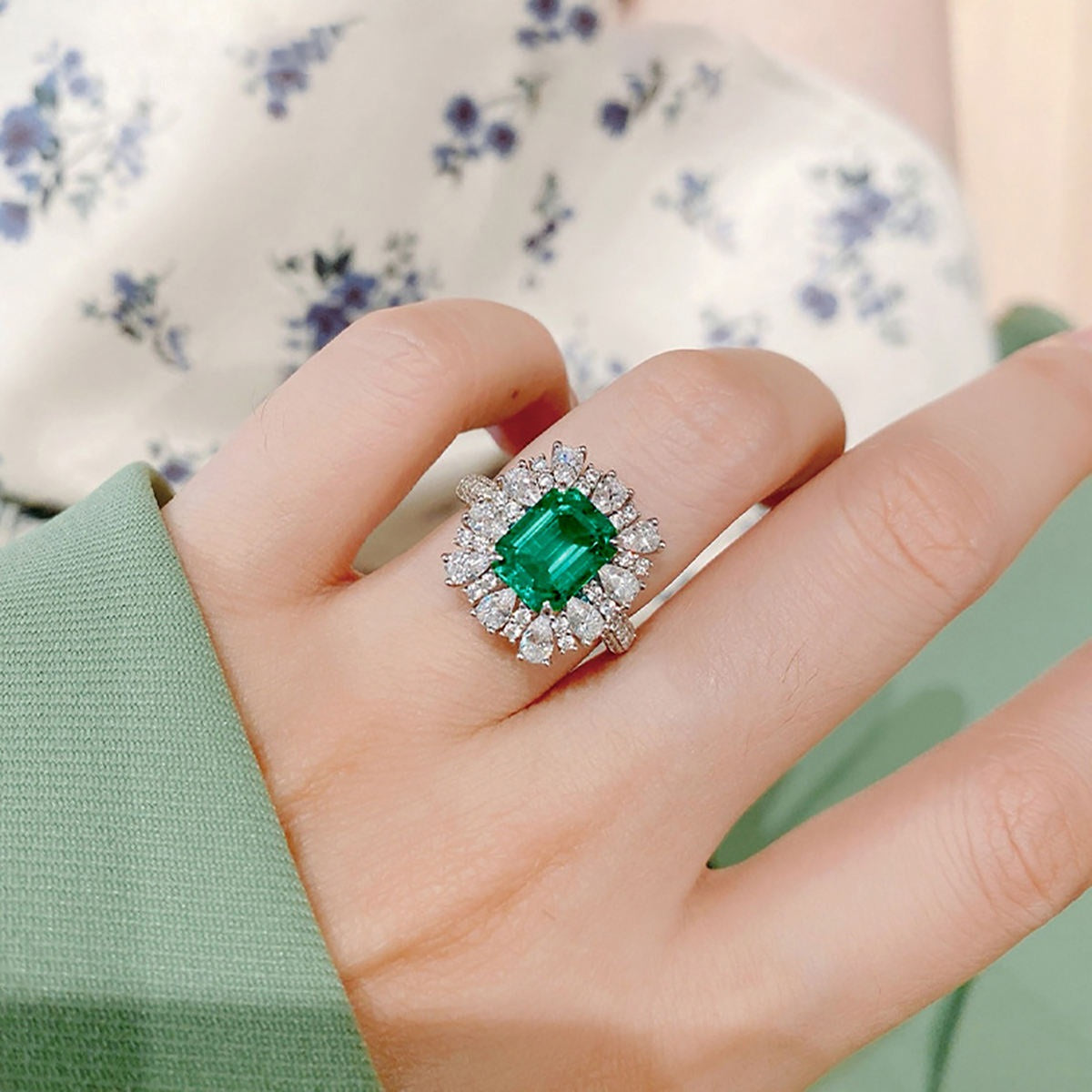 A close-up of a hand wearing a silver ring with a large emerald-cut green center stone and a halo of small clear stones, set against a soft floral fabric background.