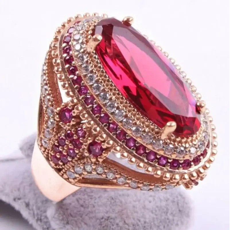 A rose gold ring featuring a large oval-cut pinkish-red central gemstone, surrounded by multiple rows of small pink and clear accent stones on a white background.