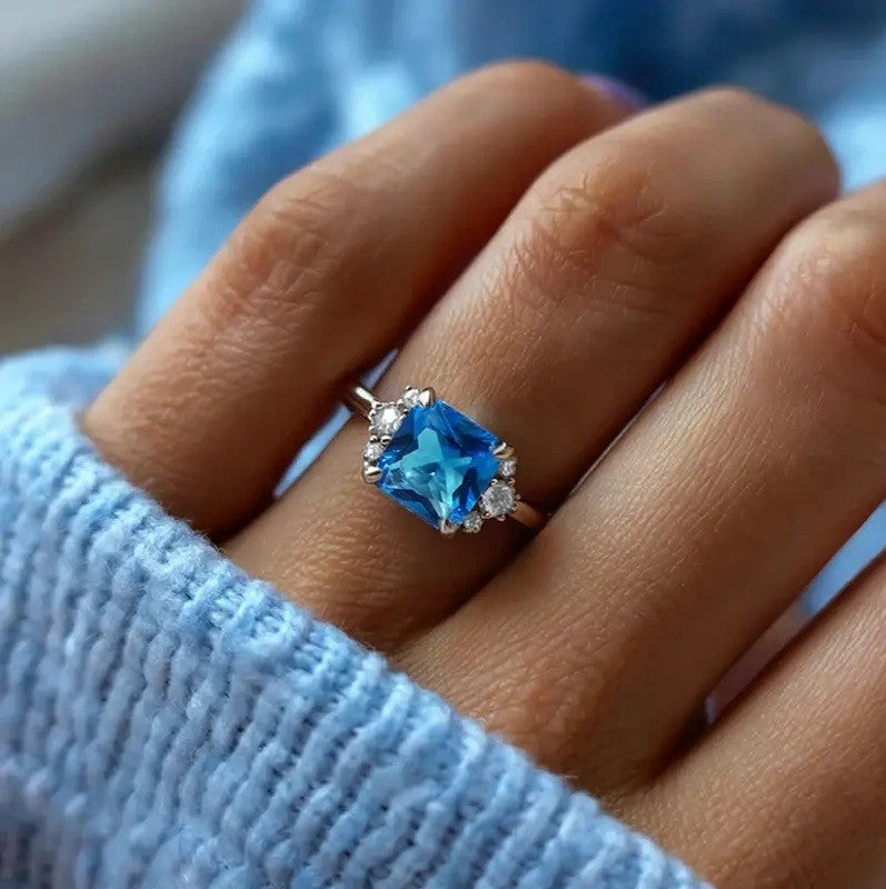 A silver ring with a cushion-cut blue gemstone and small clear accent stones, worn on a finger. The person is wearing a light blue knit sweater.