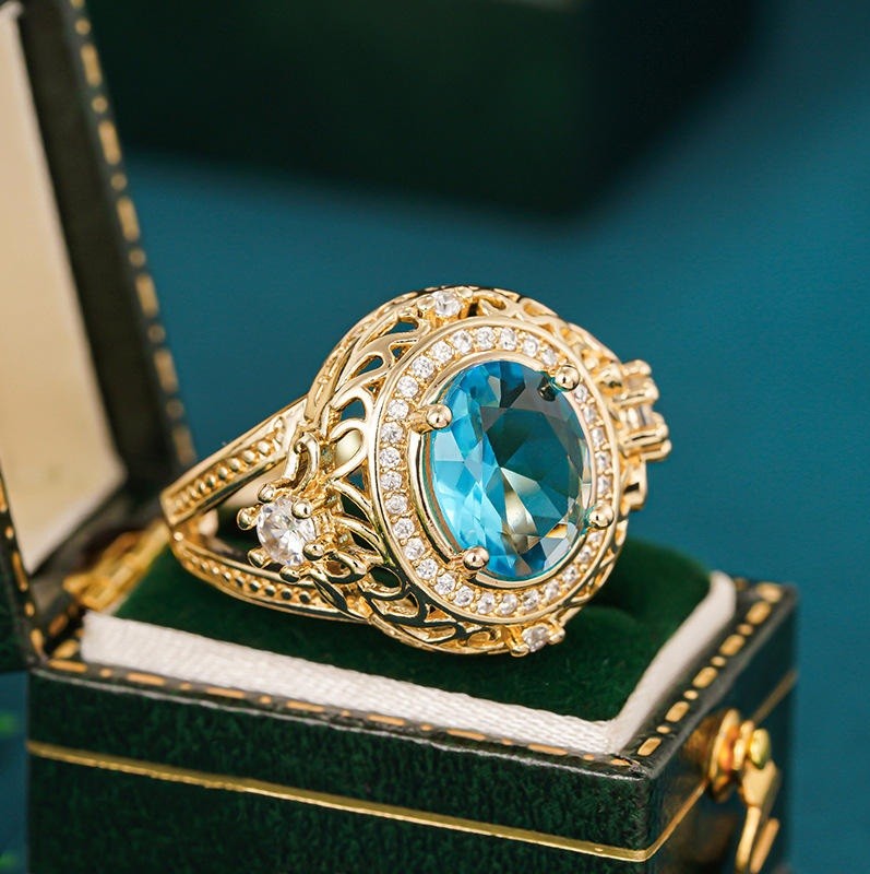 An ornate gold ring with a large oval light blue gemstone and small clear accent stones, displayed inside an open dark green jewelry box.