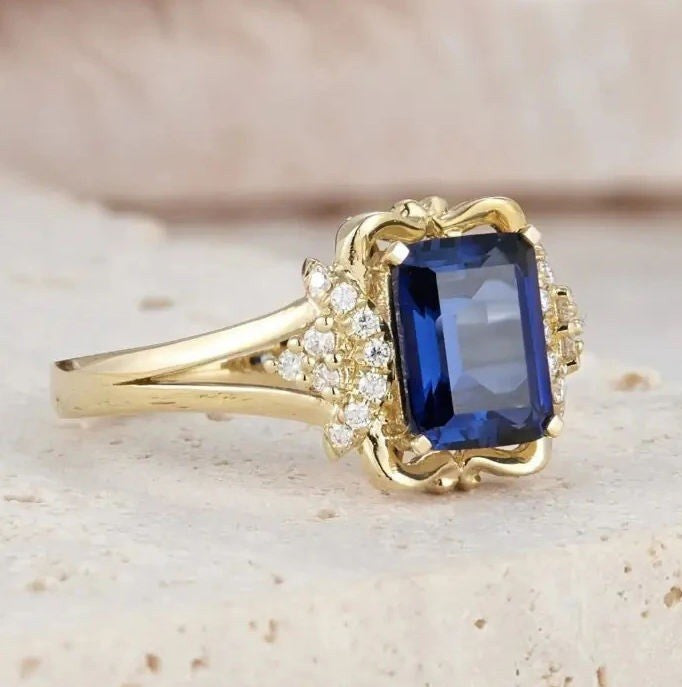 A yellow gold ring featuring a rectangular blue sapphire center stone and small clear accent stones on the shoulders, set against a light beige textured background.