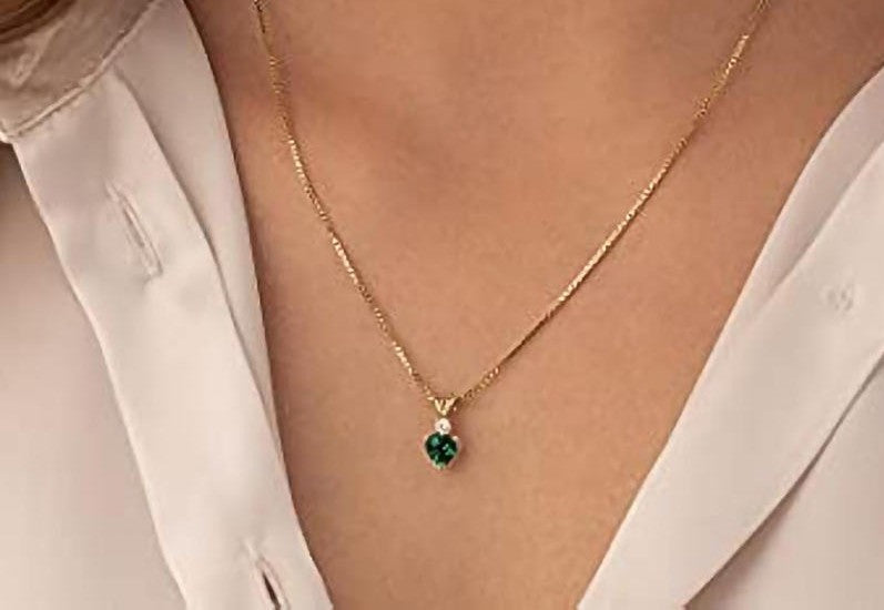 A close-up of a person wearing a delicate gold chain necklace with a small heart-shaped green gemstone pendant, styled with a white collared shirt.