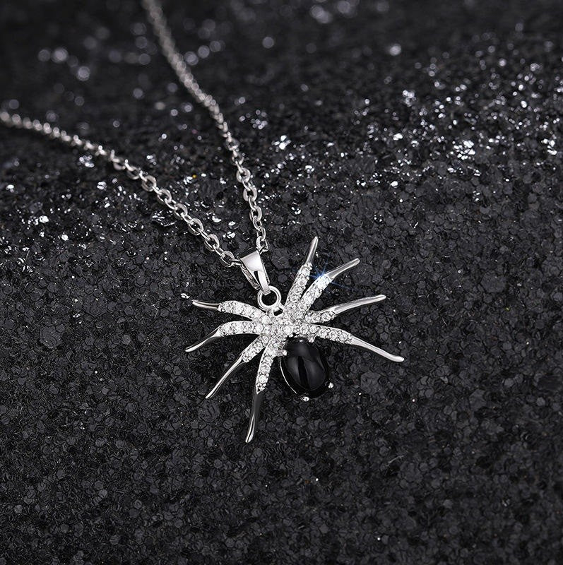 A silver spider pendant necklace with clear rhinestones and a black teardrop gemstone, displayed on a textured black background.