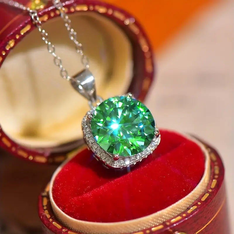 A close-up of a heart-shaped green gemstone pendant in a silver setting, resting on a red velvet jewelry box.