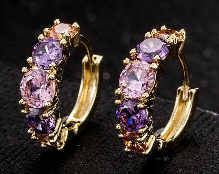 A pair of gold hoop earrings with three oval-cut purple gemstones in each earring, displayed on a textured black surface.
