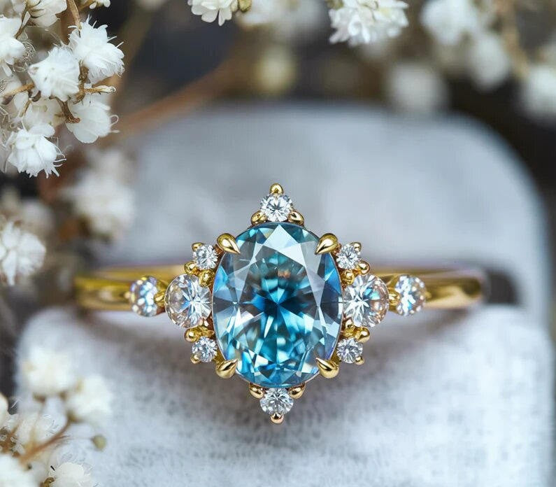 A yellow gold ring featuring a large oval-cut light blue gemstone, surrounded by small clear accent stones, displayed on a soft white ring stand with blurred white flowers in the background.