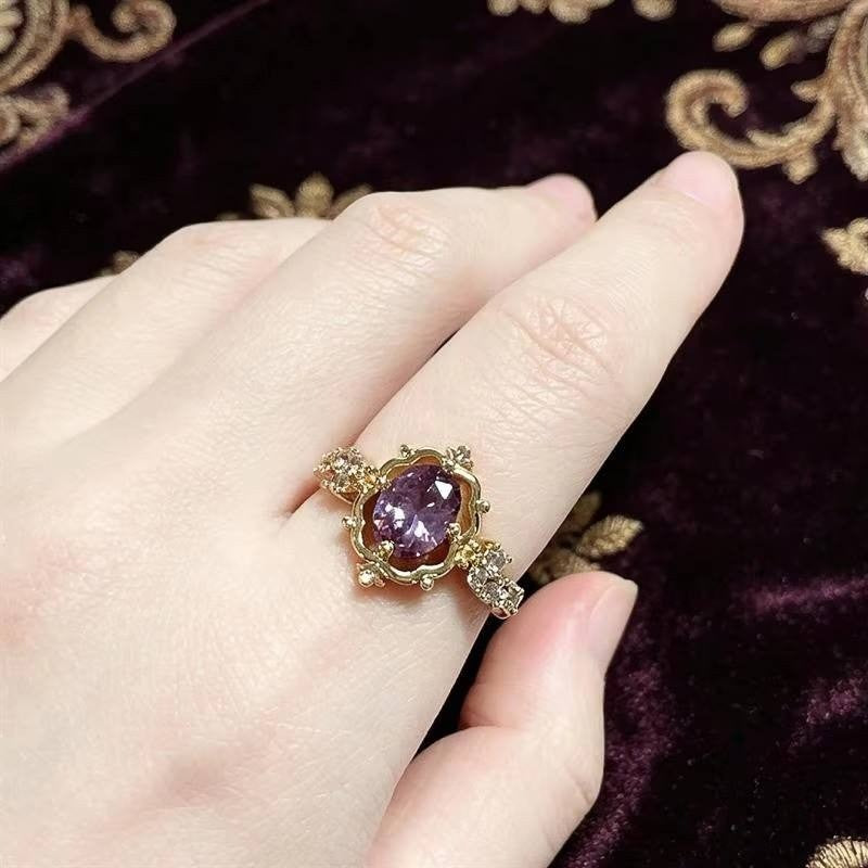 A gold ring with a central purple gemstone and small clear accent stones, shown worn on a hand against a dark purple velvet background with gold embroidery.