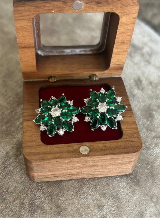 A pair of green and silver floral stud earrings displayed in an open wooden jewelry box with a red velvet lining, set against a gray textured background.