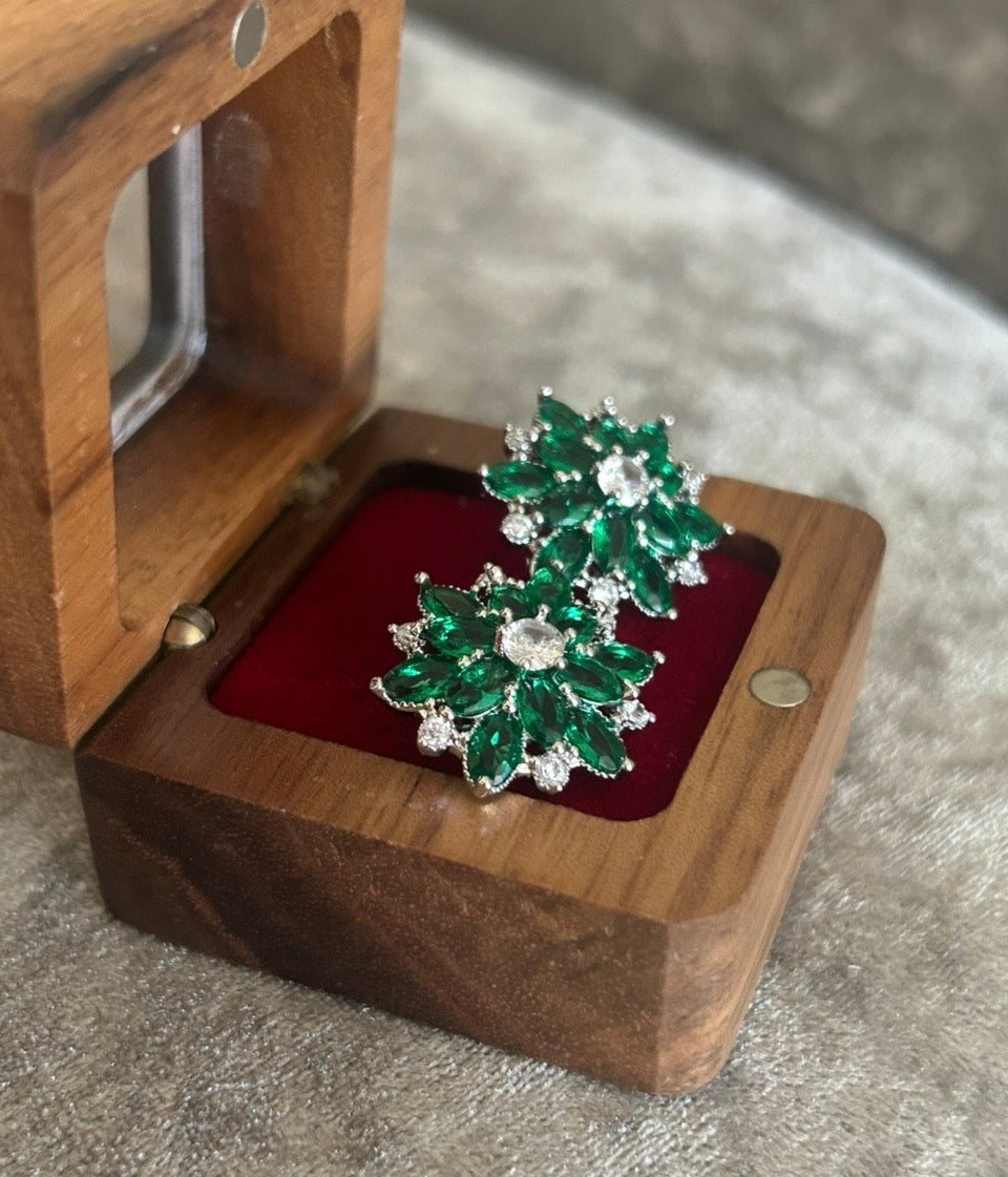 A pair of silver-toned floral earrings with green marquise-cut stones and clear round stones, displayed on a red velvet insert inside an open wooden box.