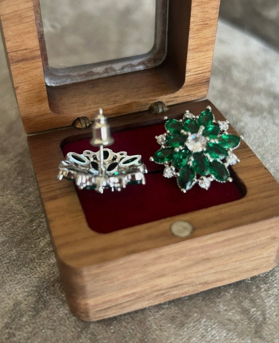 A pair of silver-toned earrings displayed in an open wooden jewelry box with a red velvet insert. One earring is silver filigree with small green stones, and the other is a green flower-shaped cluster with a clear center stone.