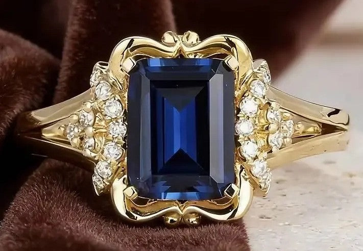A yellow gold ring featuring a large rectangular deep blue gemstone, accented by small clear stones on the shoulders, displayed on a brown textured fabric background.