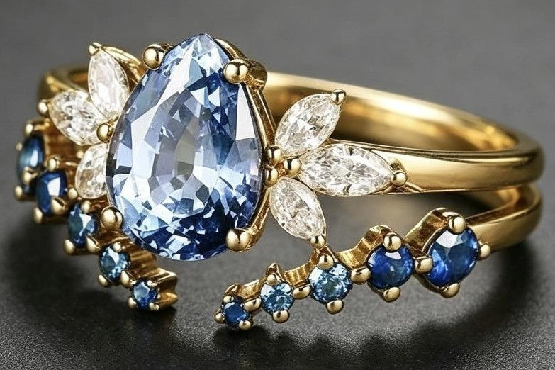A gold ring featuring a large pear-shaped light blue gemstone in the center, surrounded by small clear marquise-cut stones and smaller round blue gemstones, set against a dark gray background.