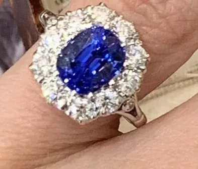 A close-up of a ring worn on a finger, featuring a large oval-cut deep blue center stone surrounded by a halo of small clear stones.