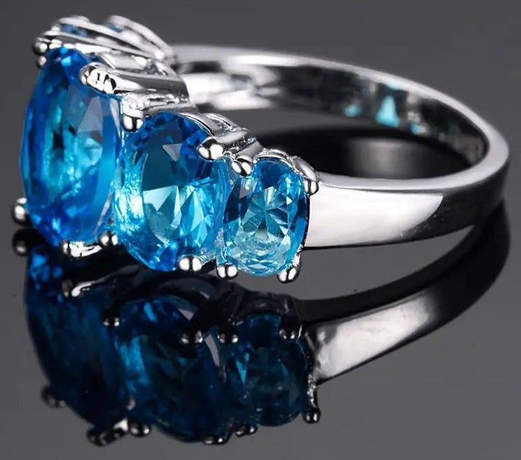A silver three-stone ring featuring oval-cut bright blue gemstones, displayed on a reflective dark gray surface.