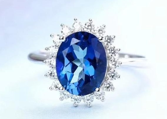 An oval-cut blue gemstone ring with a halo of small clear stones, set on a silver-toned band against a light blue background.
