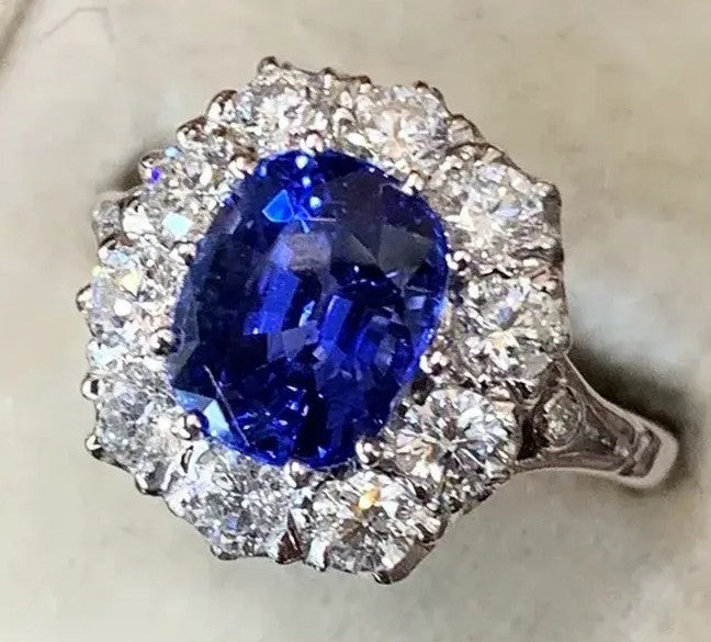 An oval-cut blue sapphire ring surrounded by a halo of round-cut clear diamonds in a silver-colored setting, shown on a light beige textured background.