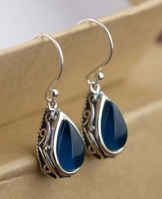 A pair of silver-toned teardrop dangle earrings with ornate filigree sides and deep blue teardrop-shaped stones, displayed on a beige jewelry stand.