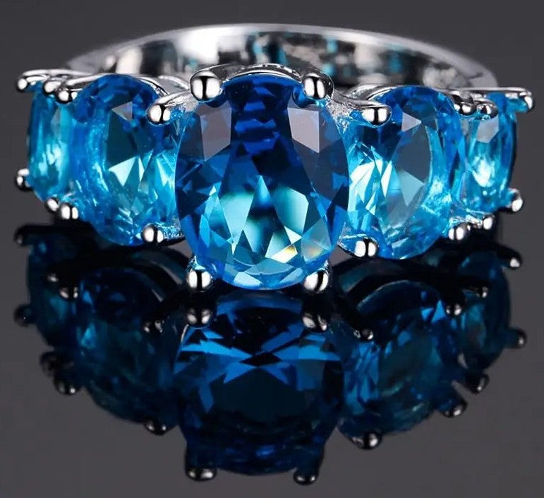 A silver ring featuring five oval-cut bright blue gemstones in a prong setting, displayed on a reflective dark surface.