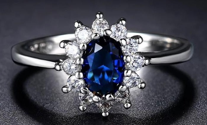 A silver ring featuring a large oval-cut deep blue sapphire center stone surrounded by a halo of small clear round-cut diamonds, set against a dark textured background.