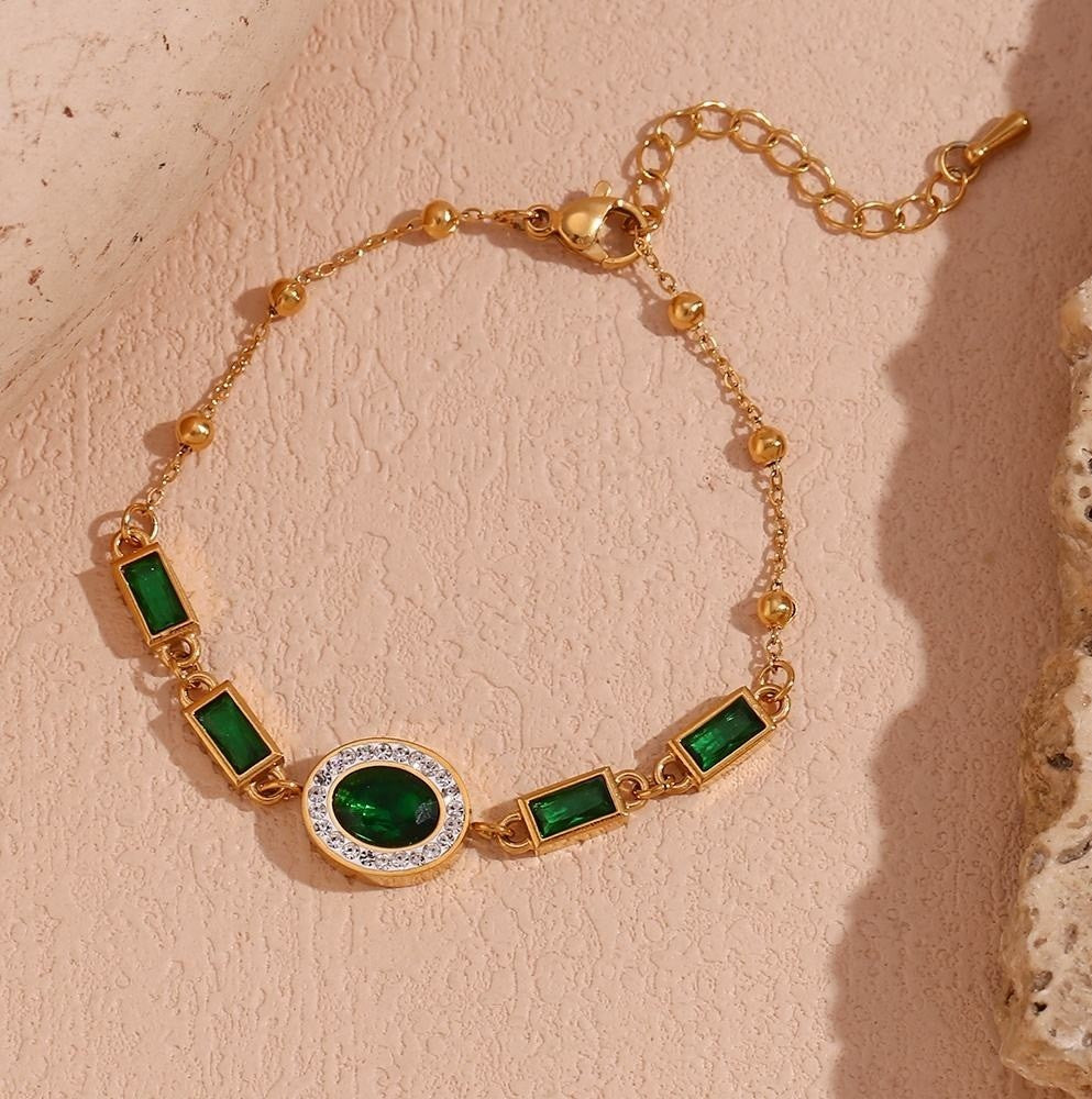 Gold chain bracelet with green rectangular and oval gemstones, a central oval green stone surrounded by clear stones, and a textured beige background.