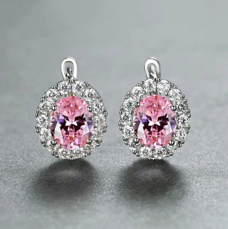A pair of oval stud earrings featuring large pink center stones surrounded by small clear stones in silver-toned settings, photographed on a smooth gray surface.