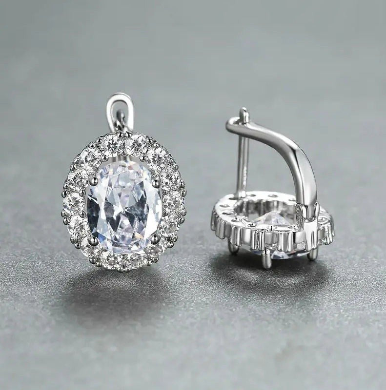 A pair of silver-toned halo earrings featuring a large oval clear gemstone surrounded by smaller sparkling stones, shown on a neutral gray background. One earring is facing forward to show the face and bail, while the other is turned sideways to display the post and back setting.