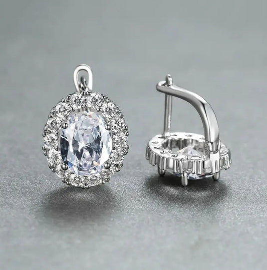 A pair of silver-toned halo earrings featuring a large oval clear gemstone surrounded by smaller sparkling stones, shown on a neutral gray background. One earring is facing forward to show the face and bail, while the other is turned sideways to display the post and back setting.