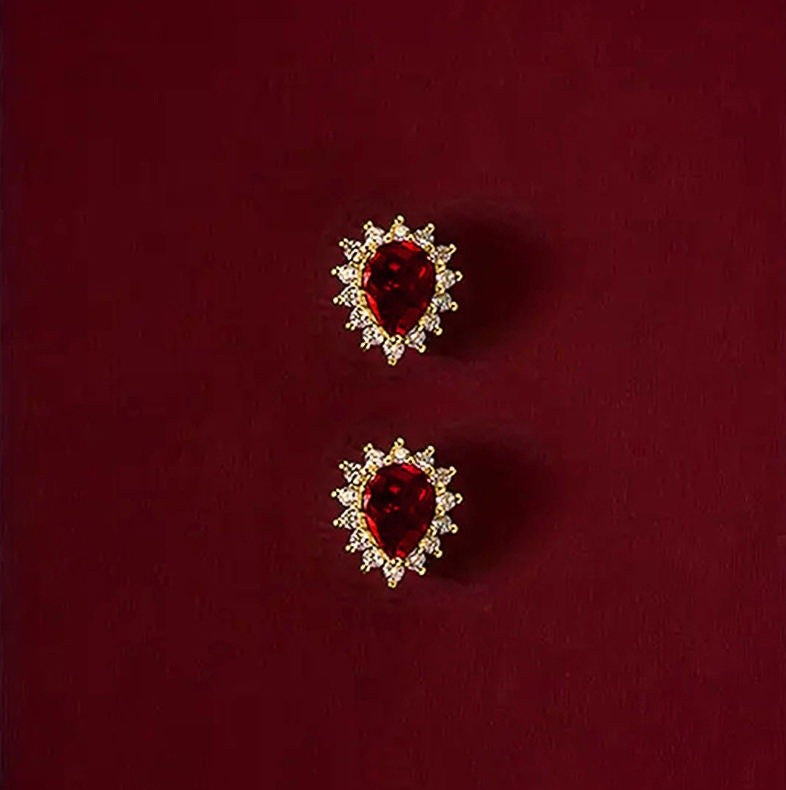 A pair of gold stud earrings featuring a central teardrop-shaped deep red gemstone 
