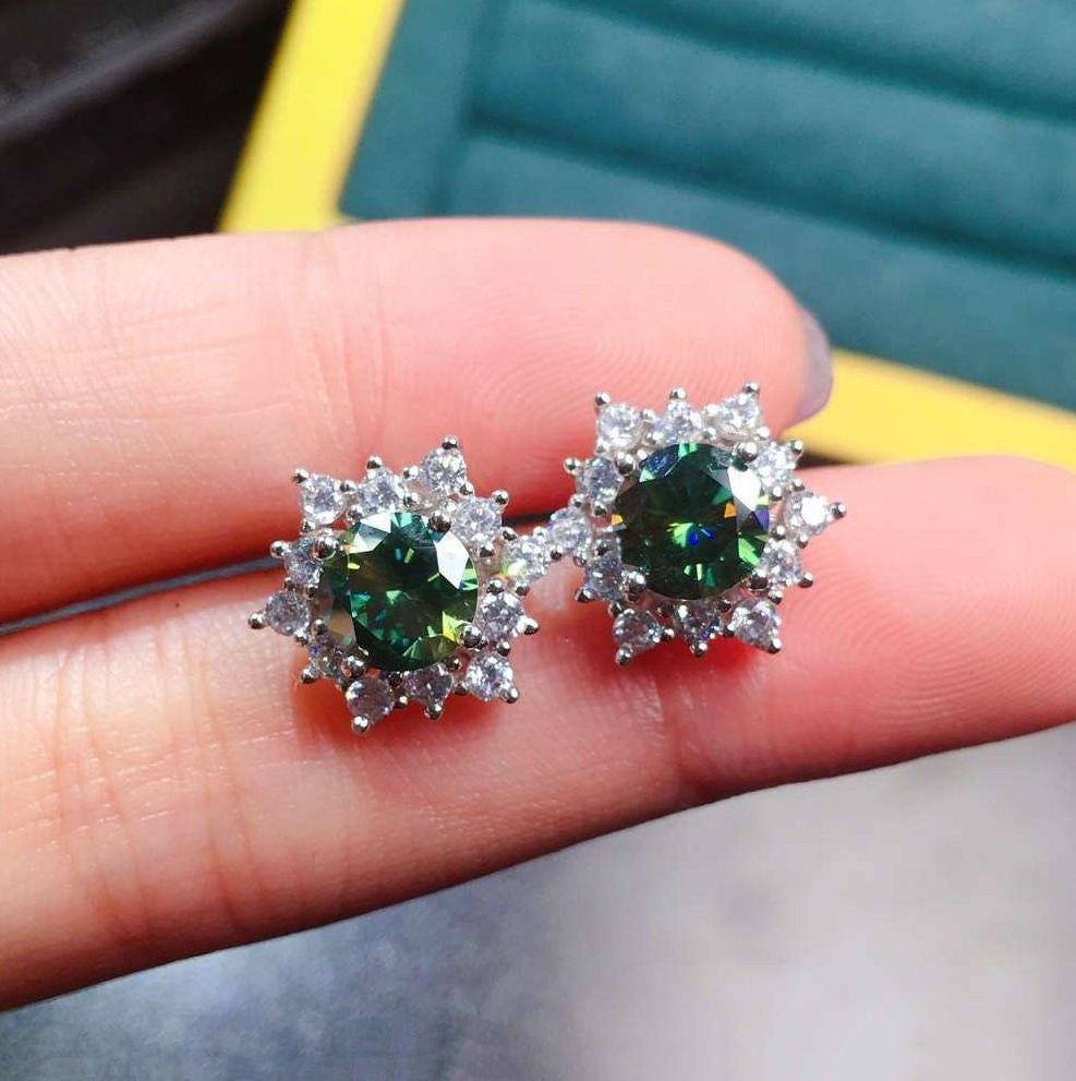 A pair of silver-toned stud earrings with oval green iridescent center stones and clear halo crystals, shown resting on a person's finger.