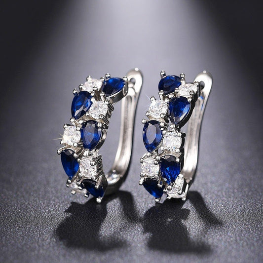 A pair of silver huggie hoop earrings featuring marquise-cut deep blue sapphires and clear round cubic zirconia stones, displayed on a dark textured surface.