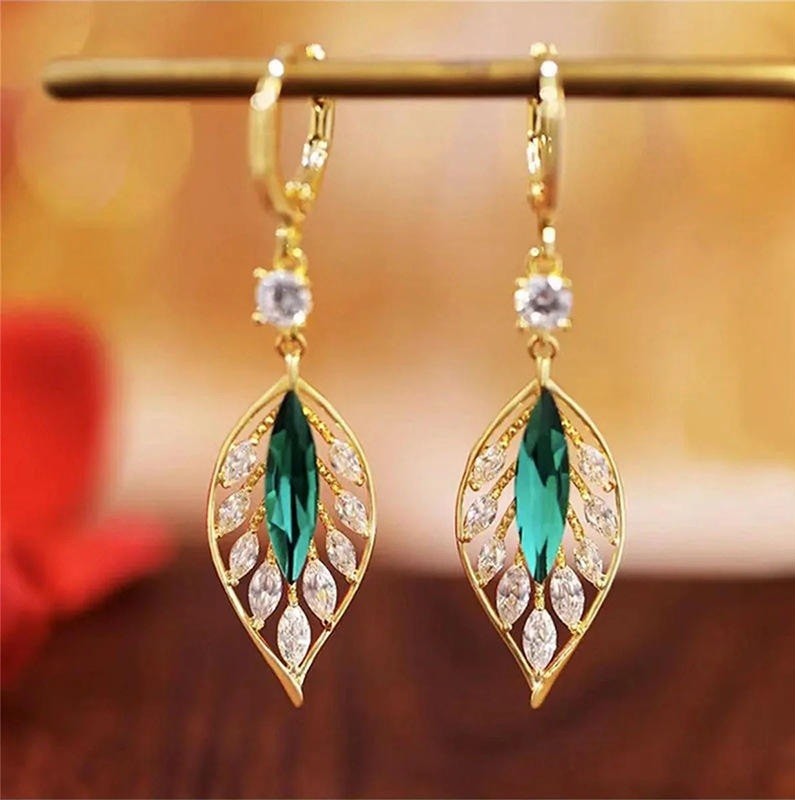A pair of gold leaf-shaped drop earrings with emerald-cut green center stones and clear rhinestone accents, hanging from a gold display bar against a warm blurred background.